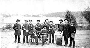Eleven men in suits and hats. Two are seated on a rough wooden bench, holding trumpets. Six are behind them, each holding a brass instrument. To the immediate right are the other three members, one standing behind a drum and one holding cymbals. About 15 feet (5 metres) behind them is a wooden fence and a faded background with trees.