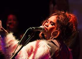 A 31 year-old woman is shown in upper body and left profile. She is singing into a microphone while raising her left arm in front of herself. She wears a khaki cloth top with pink and white woolly sleeves. Another person is blurred in the background.