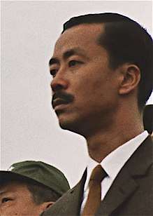  A middle-aged man with side-parted black hair and a moustache, in a black suit, white shirt and brown tie. To the left is a clean-shaven Asian man with black hair and a green military cap.