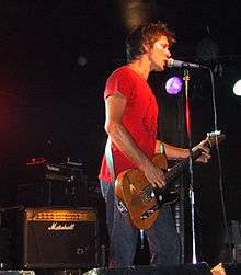 A man is shown in three-quarter side profile. He is singing into a microphone on it stand while playing a guitar. He wears a red t-shirt and jeans. Behind him are stage lights and an amplified speaker.