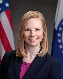 image of Nicole Galloway, smiling at the camera, wearing a collarless magenta top and dark coat, with U.S. and Missouri flags to each side in the background
