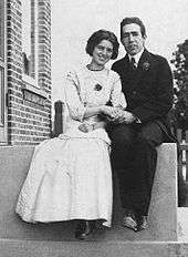 A young man in a suit and tie and a young woman in a light coloured dress sit on a stoop, holding hands