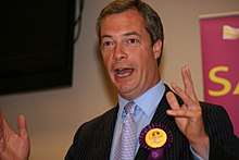 A photograph of a middle-aged white man with grey hair. His mouth is open and his arms are raised, near to his shoulders, suggesting that he is giving a public speech. He is wearing a black suit jacket with alight blue shirt and mauve tie. A purple and yellow UKIP rosette is attached to his jacket.