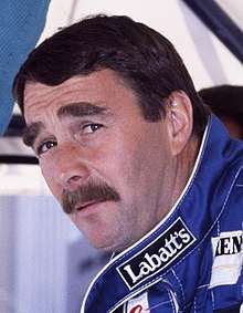 A mustached man in his late thirties is wearing blue racing overall and is looking at the camera.