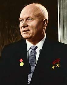 A portrait shot of an older, bald man with three medals on his chest. He is wearing a blazer over a collared shirt and tie.