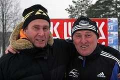 Two middle-aged men wearing winter jackets and caps embrace each other while smiling for the camera.
