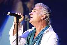 A profile shot of a man with wavy grey hair performing on stage with a black microphone. He is wearing a white shirt and a teal scarf.