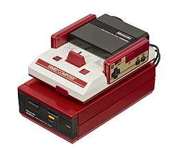 A white and red Famicom unit sits atop a candy red Famicom Disk System unit with black insertable disk drive. Two rectangular controllers, each with a D-pad and two black buttons, fit into the Famicom.