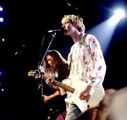 Two members of a rock band, Nirvana, pictured onstage at a show for the MTV Video Music Awards. A male singer and guitarist, Kurt Cobain, is playing electric guitar and singing into a microphone.