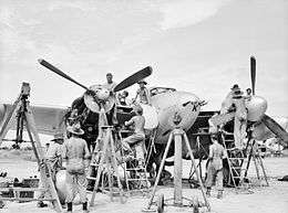 Black and white photo of a group of men conducting maintenance on a twin-engined propeller aircraft while it's on the ground. Ladders and scaffolding have been set up around the aircraft.