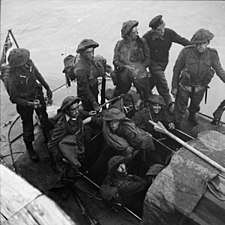 A black and white photograph showing soldiers wearing combat equipment sitting in a small watercraft beside a pier