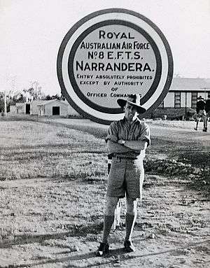 Informal portrait of uniformed man in slouch hat standing in front of sign reading "Royal Australian Air Force No. EFTS Narrandera"