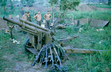 A damaged artillery piece sits in the foreground while a number of rifles resting against it. In the background a number of Caucasian soldiers stand in the background next to a hootchie.
