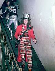 A man walks down a flight of stairs. He holds a guitar and wears a black hat studded with round metal plates. His vest and pants are chequered. His coat and shirt are plain red. His socks are striped.