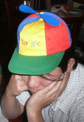 Asian man in his twenties wearing a blue, green, yellow, and red propeller hat that says "Noogle"