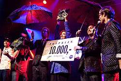 Caro Emerald, Giel Beelen and others on stage. Caro Emerald is holding a large check visibly stating 10,000,-