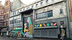 Grey stone Art Deco façade of a building on an urban street. The shutters are down at ground level and the upper windows have been replaced with window lookalikes. The ground floor is painted over with a mural containing various animals, and a crouched girl with an owl.