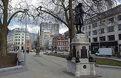 Northern end of The Centre, Bristol. A paved area with mature trees surrounded by 19th and 20th century buildings. A statue of slave trader and local philanthropist Edward Colston in the foreground, Bristol Cenotaph behind