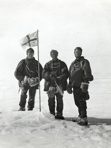  Three men stand around a flag planted in the snow.