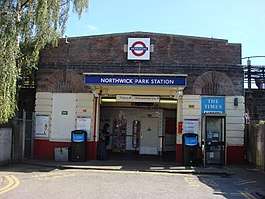 A brown-bricked building with a rectangular, dark blue sign reading "NORTHWICK PARK STATION" in white letters all under a light blue sky