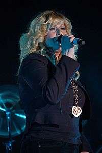 A woman with long blonde hair wearing dark clothing and singing into a microphone