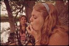 A young woman looking left with her eyes nearly closed smokes a large cigarette in a wooded setting near a body of water. Behind her another young woman looks at her