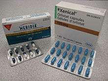 The cardboard packaging of two medications used to treat obesity. Orlistat is shown above under the brand name Xenical in a white package with Roche branding. Sibutramine is below under the brand name Meridia. Orlistat is also available as Alli in the United Kingdom. The A of the Abbott Laboratories logo is on the bottom half of the package.