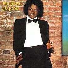 A smiling adult African American male (Michael Jackson) with a black afro, wearing a black tuxedo, white shirt, and a black bow tie. Both of his thumbs are hooked into his pants pockets with his palms and fingers facing forward and splayed out. The sides of his jacket are tucked behind his hands as he leans back slightly, giving a playful, casual touch to his formal look. Behind him there is a brown brick wall and to the side of his head are "MICHAEL JACKSON" in yellow chalk writing and "OFF THE WALL" in white chalk writing. "JACKSON" and "WALL" are separately underlined.
