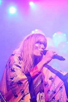 Oh Land is performing live into a microphone while wearing a kimono and glitter is spread across her face.