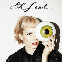 A color photograph of a female singer with a bob haircut holds a large green eyeball against her face. "Oh Land" in cursive appears above the woman.