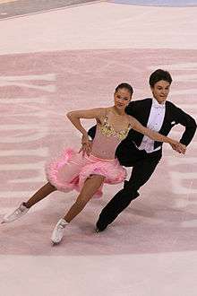 A man and woman ice dancing; the woman, on the left, is wearing a light-pink knee-length dress with a flowing skirt, and the man is wearing a classic tuxedo with a white shirt, cummerbund, and bow-tie