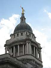 The stone dome at the top of a building, columns below, with a bronze statue on top depicting a woman holding a sword in one hand and a pair of scales in the other
