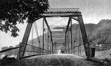 A black and white image taken looking along the bridge, with a high hill in the background. Signs visible hanging from the bridge superstructure say that it is a toll bridge and there is a $10 fine for crossing the bridge at a speed faster than a walk