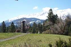Photograph of Old Chief Joseph's gravesite, with a stone pillar on top of a hillock and the Wallowa Mountains rising in the background.