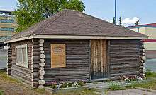 A small one-story square brown building of logs with the ends unfinished at the corners. It has a peaked roof with brown shingles and yellow trim on top. In front are flower beds; it has a yellow sign with fancy text headed "Yellowknife's Original School" at left and a wooden door of vertical boards in the middle. Behind it is a tree and part of two larger modern buildings.