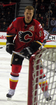A man with short hair stares forward. He is in full hockey gear and wearing a red uniform with yellow, white and black trim and a stylized "C" logo on the chest