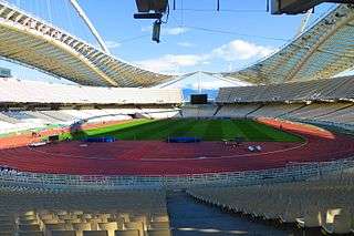 An orange oval track with a grass infield, with grass cut longitudinally, with a shadow cast over most of the venue