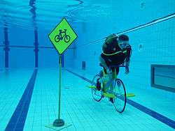 A cyclist wearing a scuba tank, fully submerged in a pool, pedals past a bicycle warning road sign.