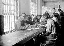 Black and white image of several prisoners, mostly of African heritage, sitting at a desk and writing. There are bars on the windows.