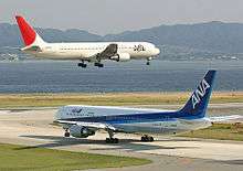 A white and red-tailed Japan Airlines aircraft above runway, with landing gears down, and an All Nippon Airways in blue and white livery taxiing