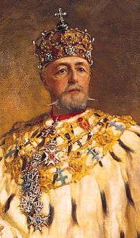 An elderly, bearded man in ornate robes, wearing a jewelled crown surmounted by a cross, looks straight out of the picture.