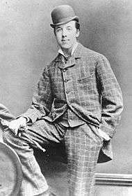 Oscar Wilde posing for a photograph, looking at the camera. He is wearing a checked suit and a bowler hat. His right foot is resting on a knee high bench, and his right hand, holding gloves, is on it. The left hand is in the pocket.