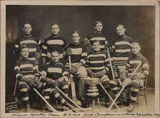 Two rows of men in striped hockey uniforms, the first row seated. In front of them on the floor is a large trophy