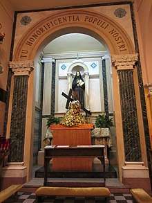 Kneeler in front of a statue holding a cross while clothed in red and gold garments. Behind it is a statue of a nun, all beneath a rounded arch