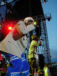 Two black men rap into microphones whilst on stage. One wears a blonde wig, a grey jacket and blue trousers, and the other wears a green chequered hat, white shirt and khaki shorts.