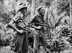 Two men wearing military style uniforms, one wearing a slouch hat, the other a general service cap, carrying weapons in a jungle setting