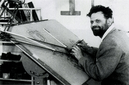 A bearded man in his mid-30s, dressed in a shirt and woolen pullover, is sitting at a drawing board. His pencil is poised in his right hand, and he is smiling off to the left of the camera.