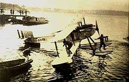 Single-engined biplane with floats, parked on water, with one crewman on float and another on lower wing
