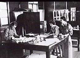 Three women in overalls around a large desk