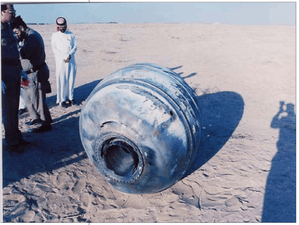 Cylindrical rocket fragment on sand, with men looking at it